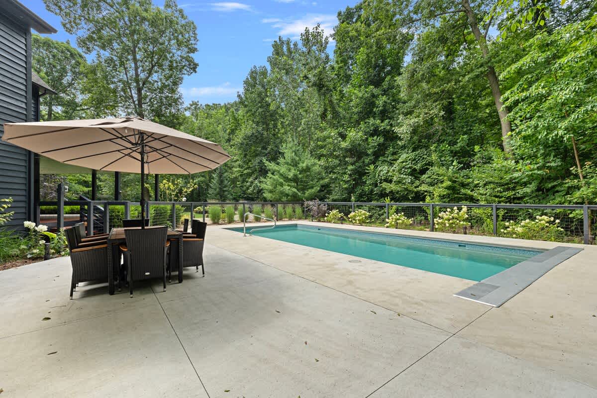 A long, rectangular outdoor swimming pool is set into a spacious concrete patio, flanked by an outdoor dining area with an umbrella and surrounded by lush green trees and landscaping.