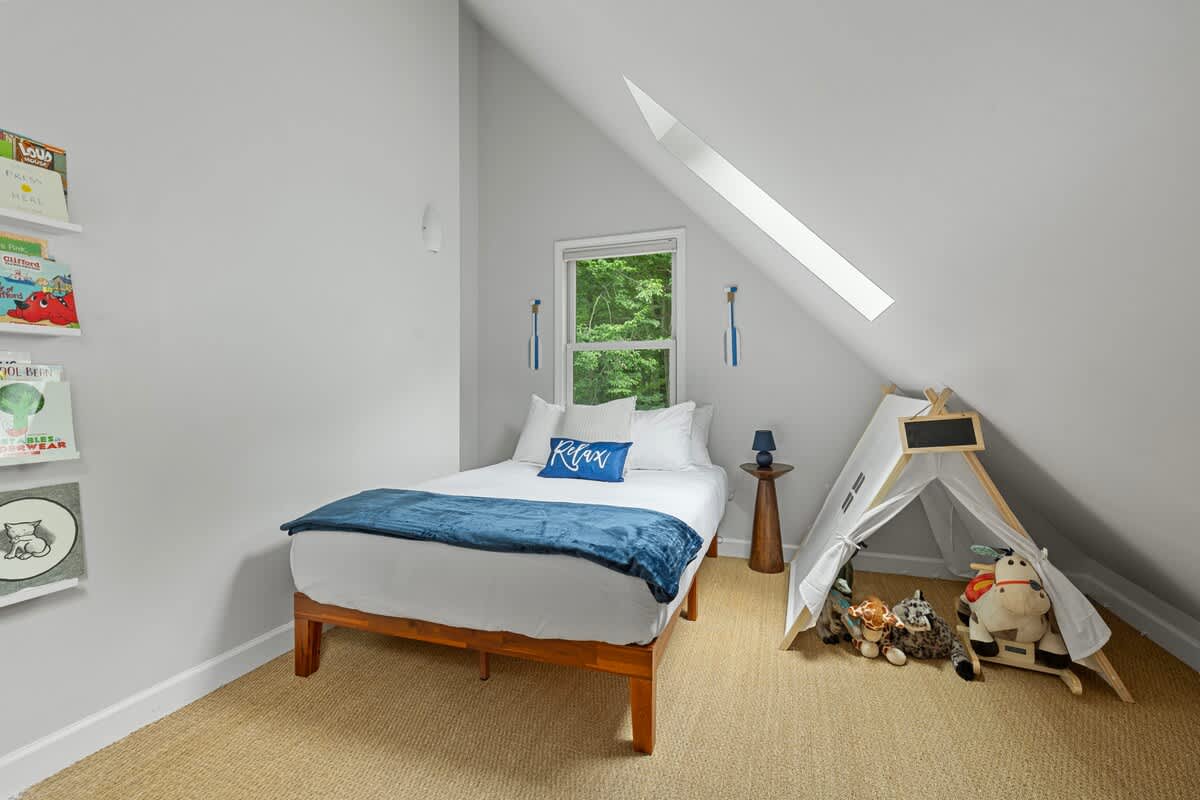 A charming children's bedroom featuring a single bed with a blue throw, a white teepee with toys, and a sloped ceiling with a skylight. Bookshelves line the wall, on a natural fiber rug.
