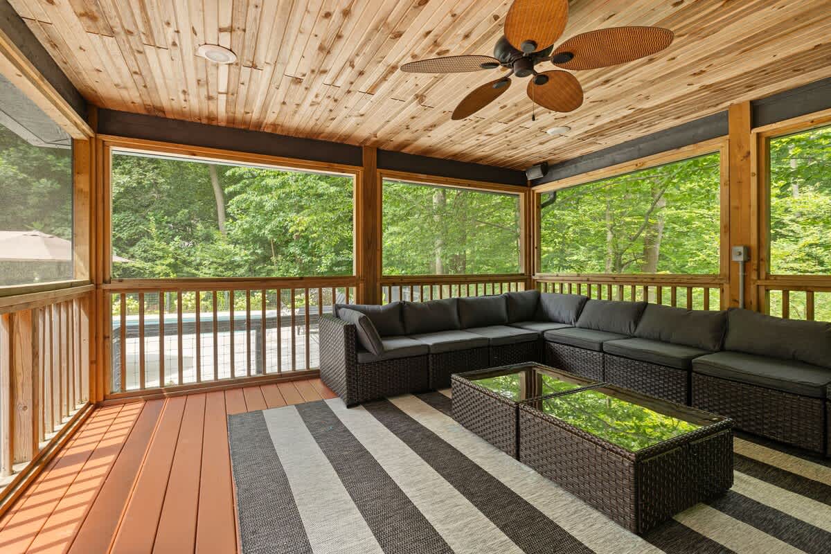 Screened-in porch with a large dark wicker sectional sofa and glass-top coffee tables on a striped rug, featuring a wood-paneled ceiling with a fan and views of the surrounding trees.