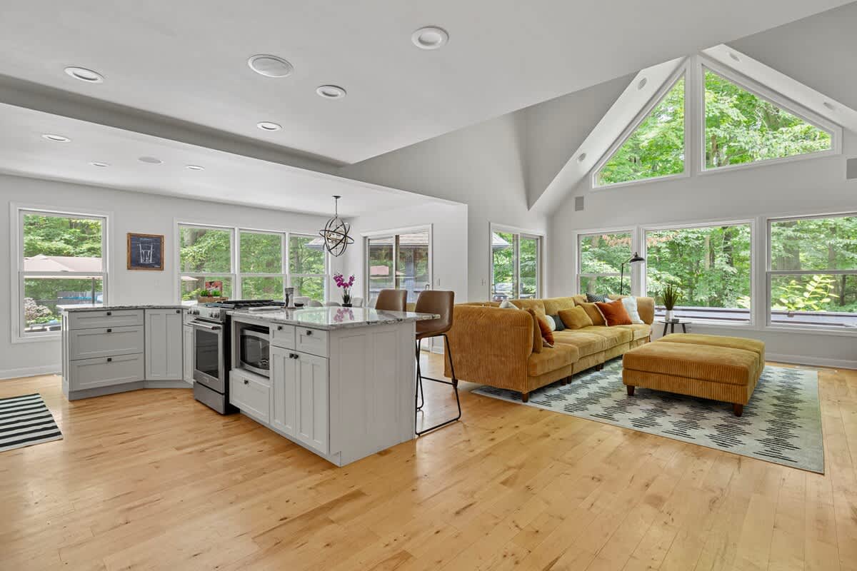 Expansive open-concept living area with light hardwood floors, a large mustard yellow sectional sofa, and a modern kitchen island, all bathed in natural light from numerous windows overlooking lush greenery.