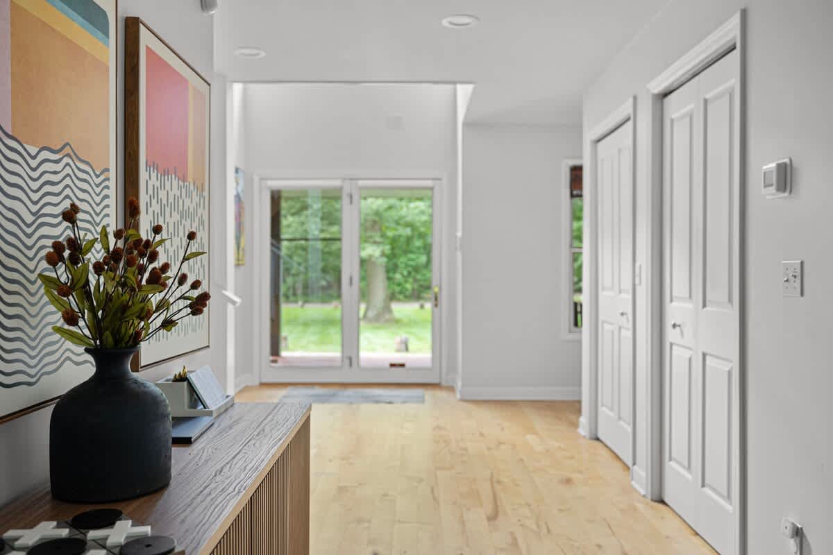 Bright foyer with light hardwood floors, white walls, and a console table with decor, leading to a glass double door that opens to a green outdoor space.
