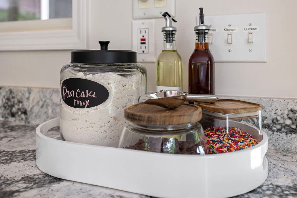 A white oval serving tray on a granite countertop holds a glass jar of "Pancake mix," chocolate chips, and colorful sprinkles, alongside bottles of cooking oil or syrup.