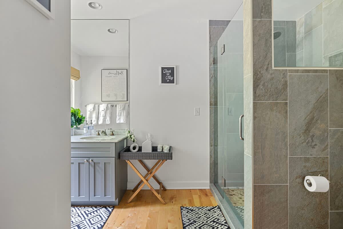 A contemporary bathroom with a spacious walk-in shower featuring gray tiled walls and a glass enclosure. A wooden stool with a tray sits next to the vanity area, with hardwood floors.
