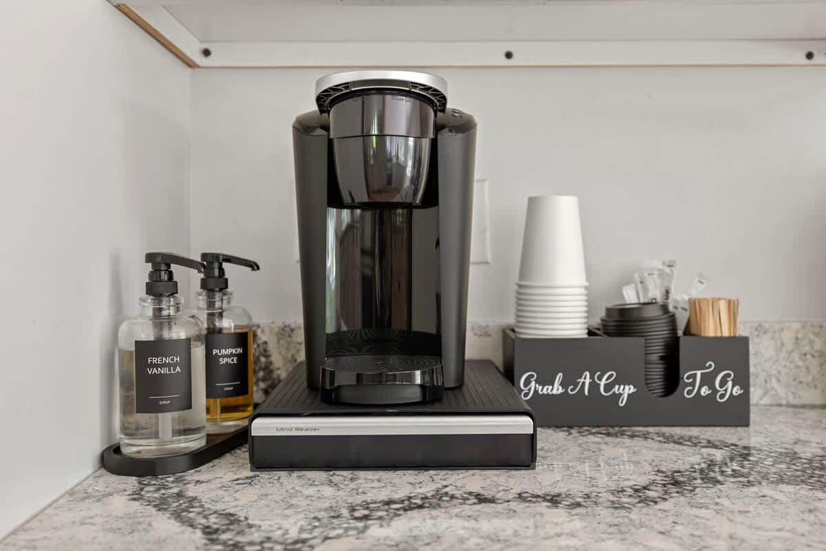 A dedicated coffee station on a speckled granite countertop, equipped with a black single-serve coffee maker, clear pump dispensers for syrups, and a "Grab A Cup To Go" caddy with disposable cups.
