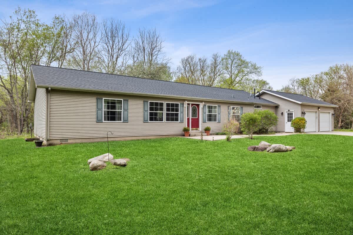 Charming single-story house with light beige siding, a red front door, and green shutters, surrounded by a well-maintained green lawn under a clear blue sky.