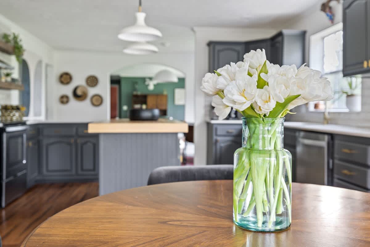 Close-up view of a round wooden dining table adorned with a vase of fresh white tulips, with the dark gray kitchen cabinets and island visible in the softly blurred background.