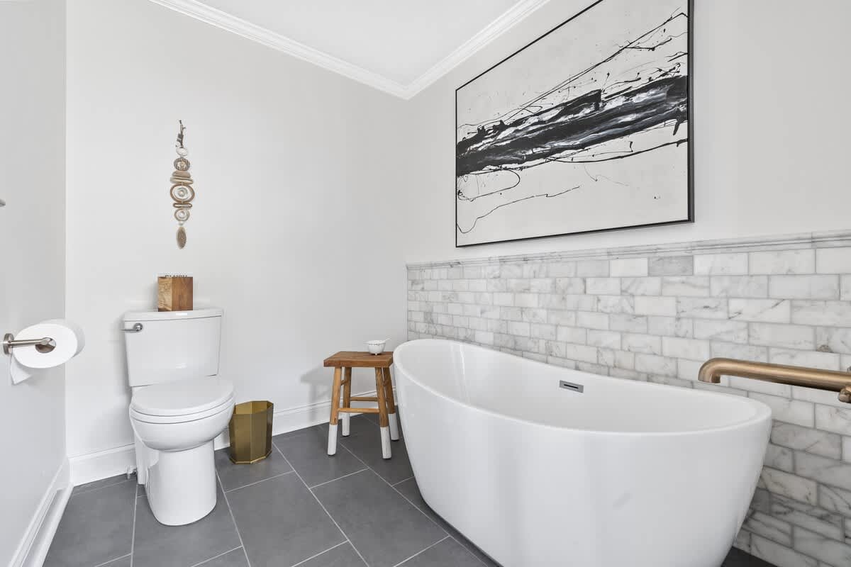 Elegant bathroom featuring a white freestanding bathtub, a toilet, and white marble subway tile wainscoting, complemented by a large abstract black and white artwork.