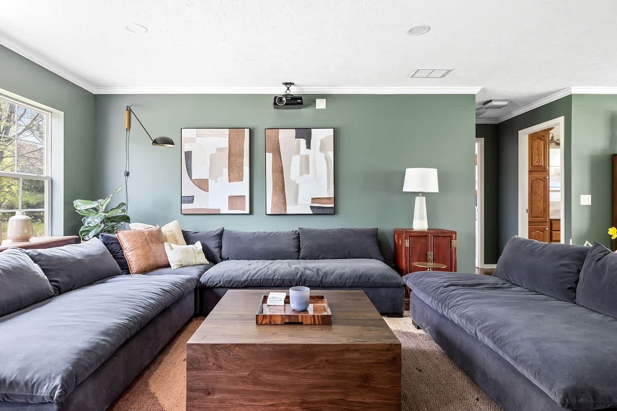 A comfortable living room with a large dark gray sectional sofa, a wooden coffee table, and abstract artwork on a sage green wall, featuring a projector and ample natural light from windows.