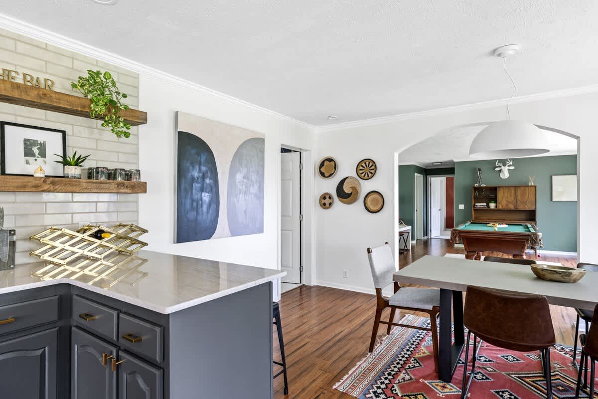 Modern kitchen with dark gray shaker cabinets, light quartz countertops, white subway tile backsplash, and open wooden shelving, looking into an open-concept dining and game room.