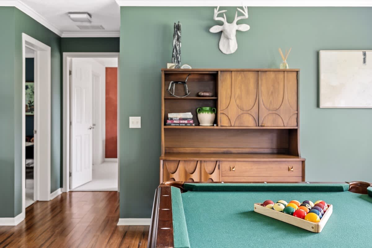 Inviting game room featuring a green-felt pool table with racked balls, a vintage wooden hutch with decor, and sage green walls, leading to a hallway with white doors.