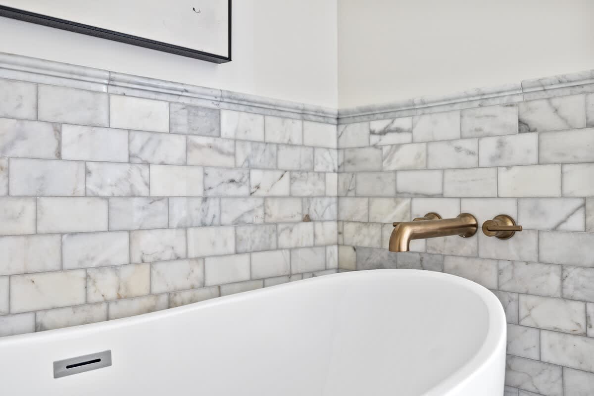 Close-up view of a contemporary white freestanding bathtub against a wall with white marble subway tile wainscoting and a wall-mounted bronze faucet.