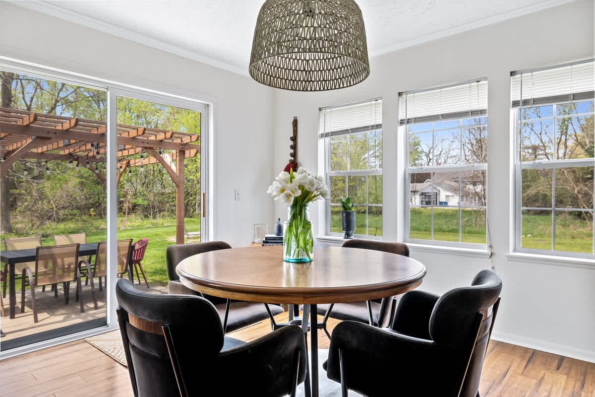 Bright dining area with a round wooden table, four upholstered chairs, and a woven pendant light, featuring a large sliding glass door and multiple windows offering expansive views of the lush green backyard.