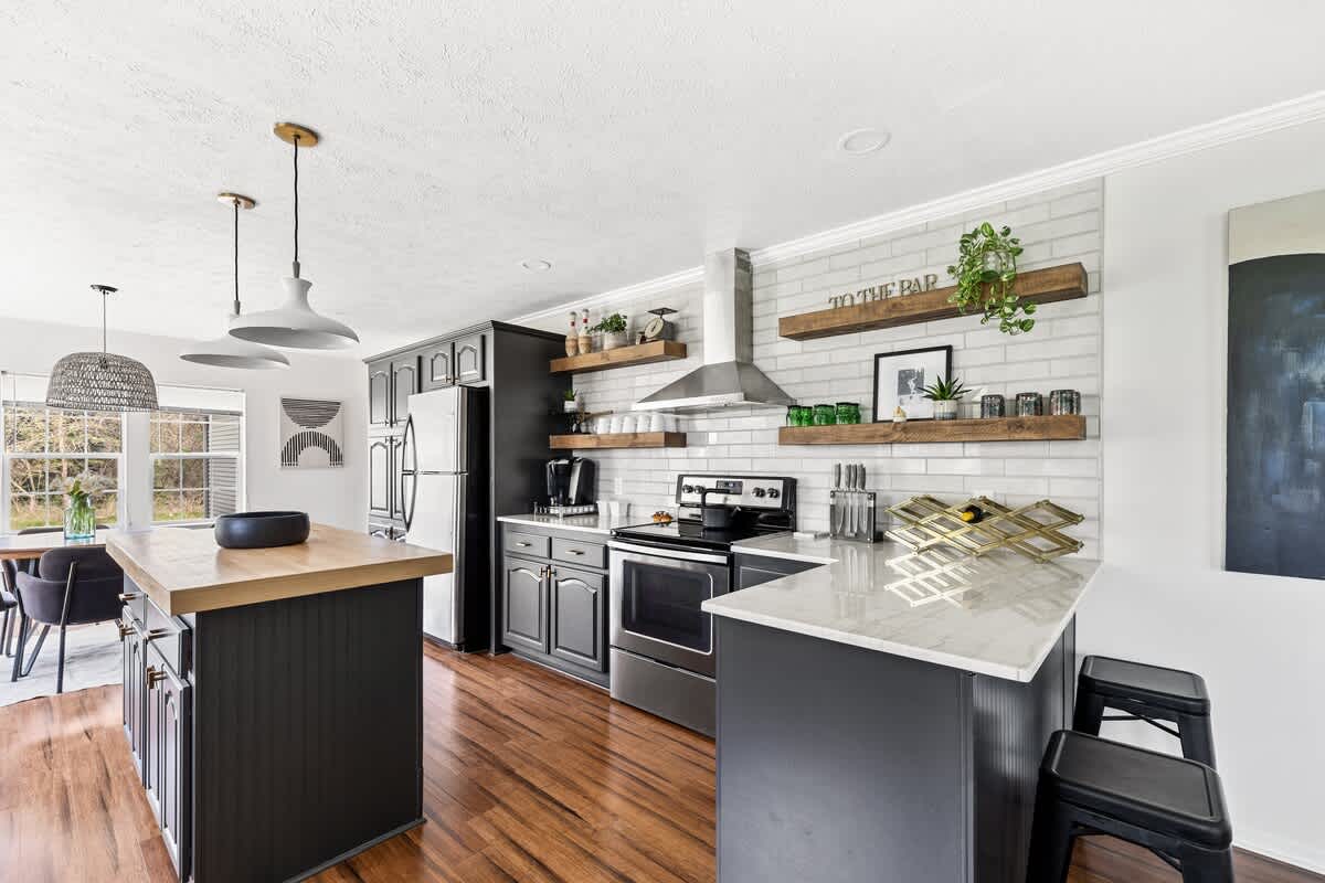 A modern kitchen with dark gray cabinets, a white subway tile backsplash, and a wooden island, extending into a dining area with a table and chairs, all on hardwood floors.