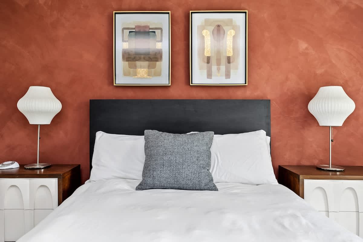 Stylish bedroom featuring a bed with a dark headboard, white bedding, two modern nightstands with unique white lamps, and an accent wall painted in a warm terracotta hue.