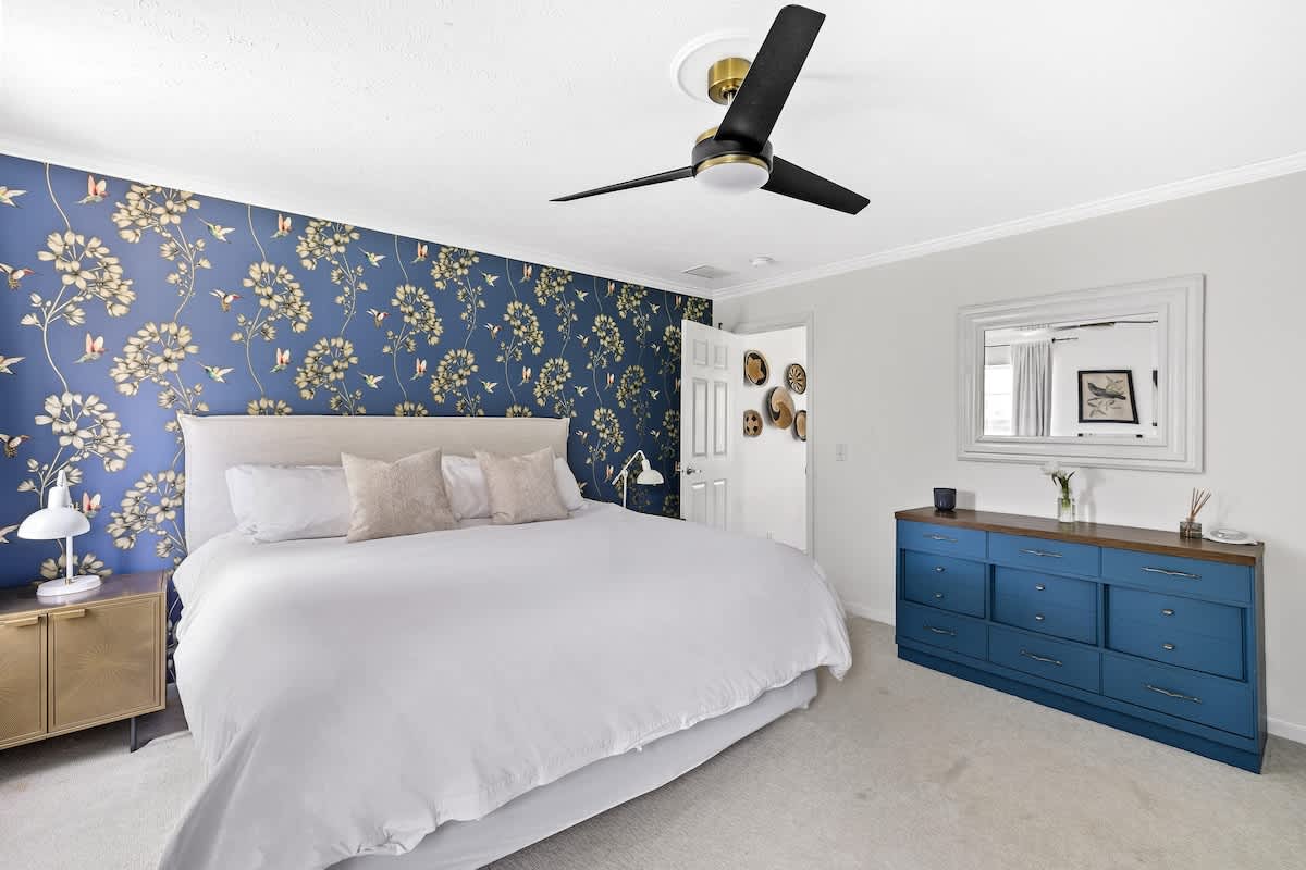 Bright and stylish bedroom featuring a large bed with a white duvet, a bold blue floral wallpaper accent wall, a long blue dresser with a wooden top, and a modern ceiling fan.
