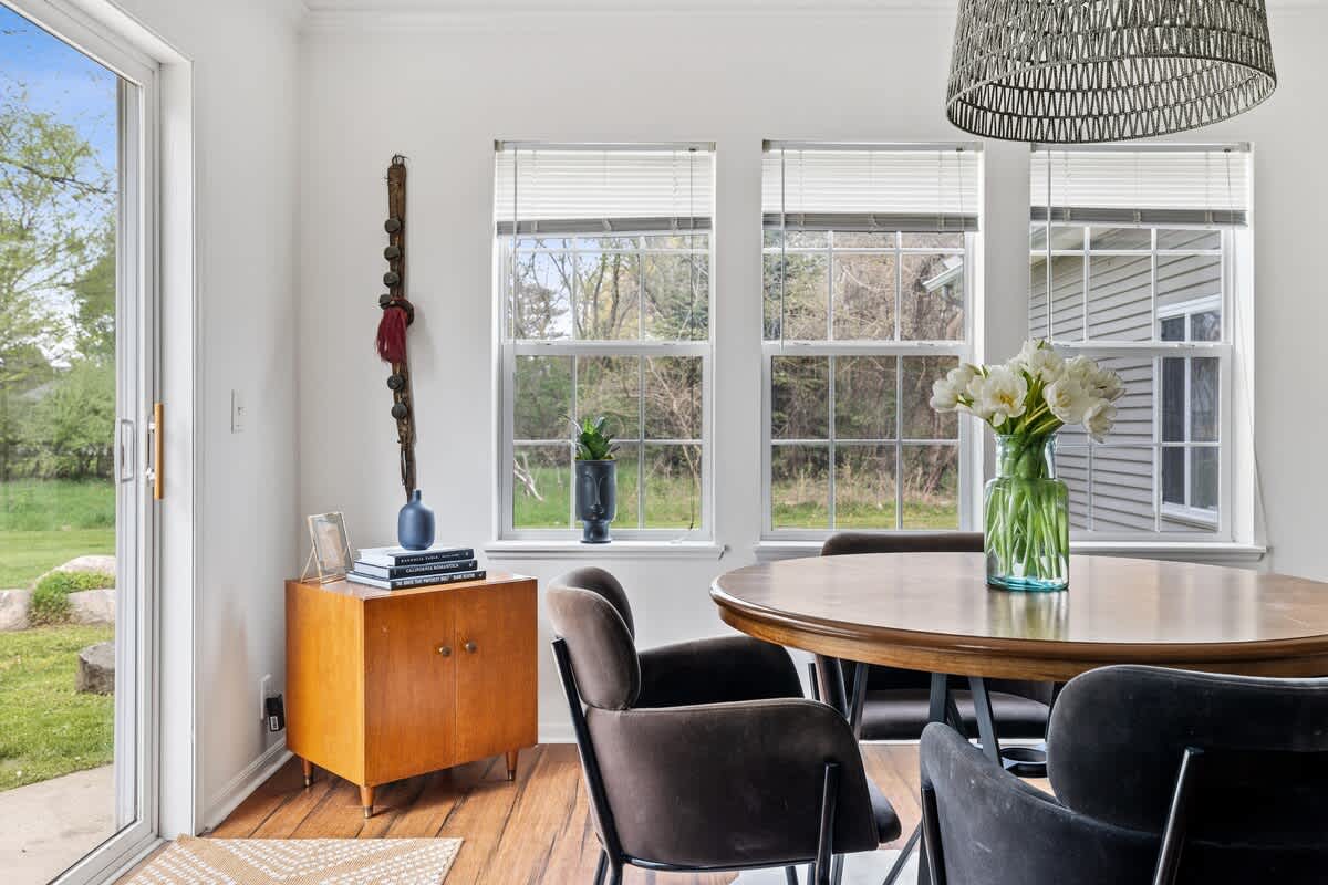 Cozy dining nook with a round wooden table, dark velvet chairs, and large windows overlooking the green backyard, complemented by a small mid-century modern wooden cabinet with decorative items.