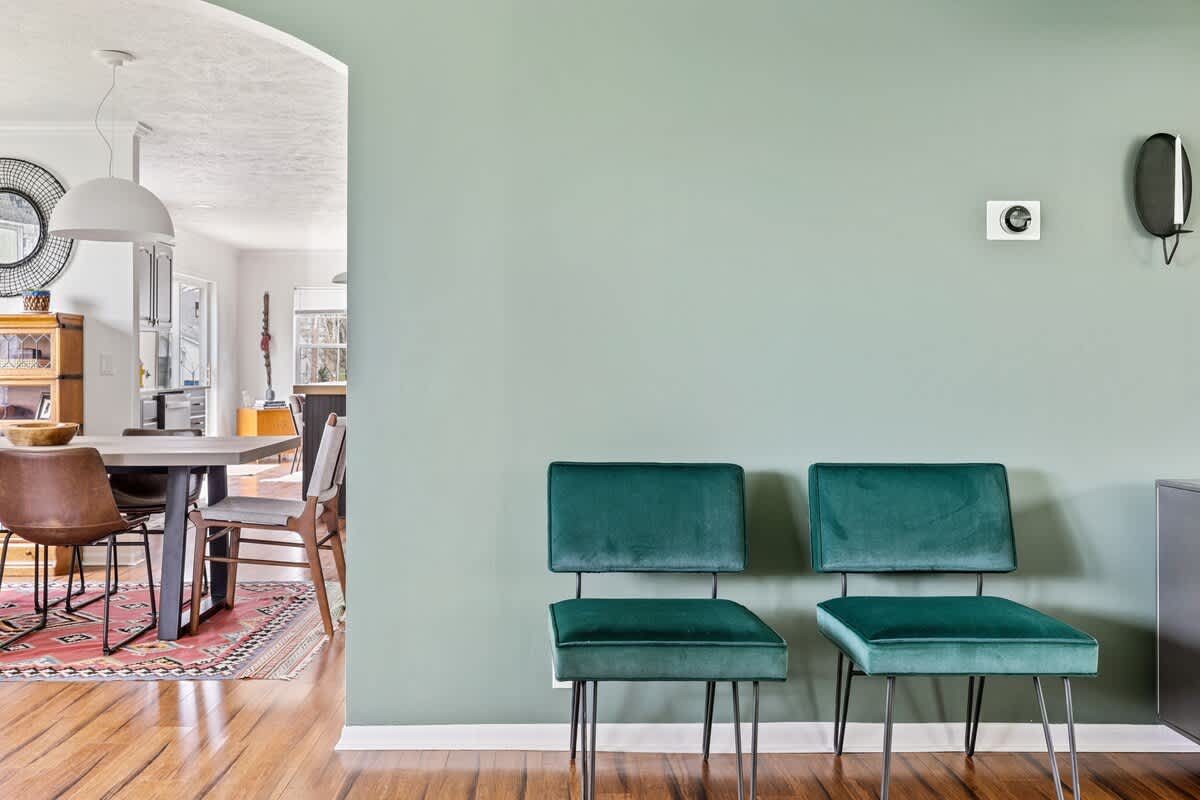 A sage green wall with two modern emerald green velvet chairs, offering a glimpse through an arched doorway into a dining area with a table and a kitchen beyond.