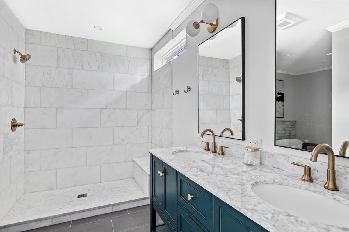Spacious walk-in shower with white marble subway tiles and a built-in bench, adjacent to a double vanity with marble countertops, brass fixtures, and dark blue cabinetry.