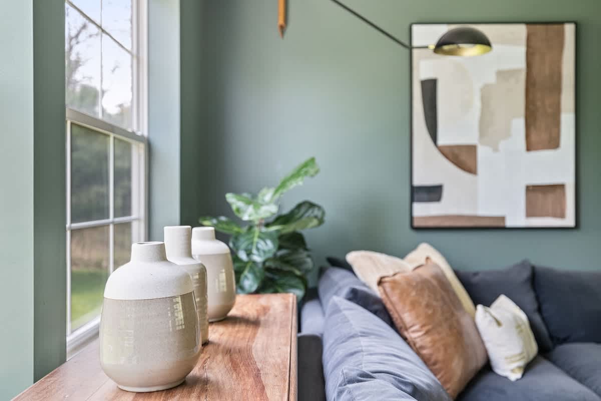 A close-up of a living room detail, showing two ceramic vases on a wooden console table next to a window, with a fiddle leaf fig plant and a dark gray sofa in the background.