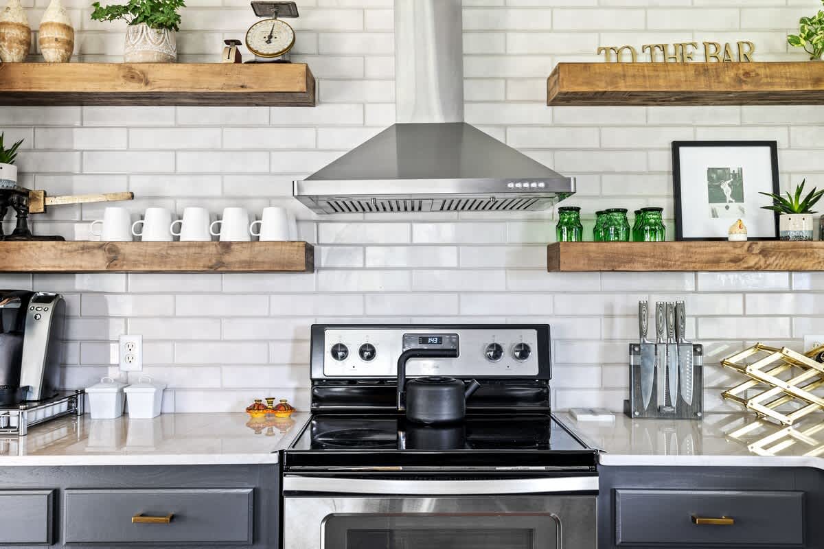 A detailed view of the kitchen stove and range hood, set against a white subway tile backsplash with open wooden shelves displaying decorative items and kitchenware.