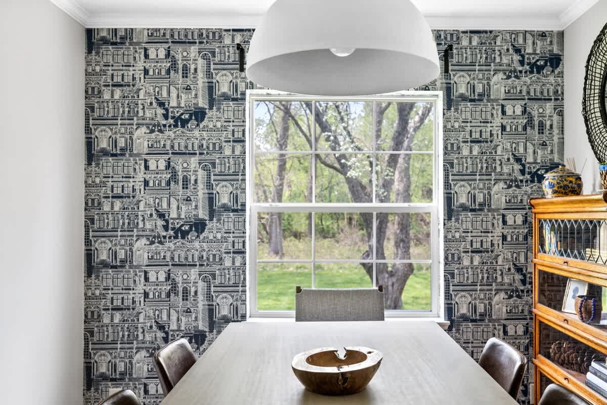 Elegant dining room with a large rectangular table, modern white pendant light, and a striking black and white architectural wallpaper accent wall, featuring a large window overlooking trees.