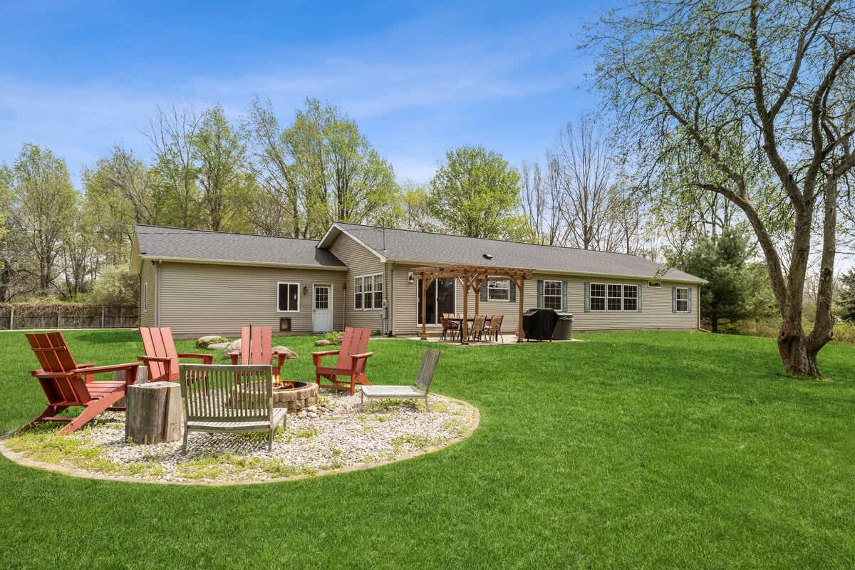 Expansive backyard featuring a light-colored ranch-style house with a covered patio, a large green lawn, and a gravel fire pit area with red Adirondack chairs.