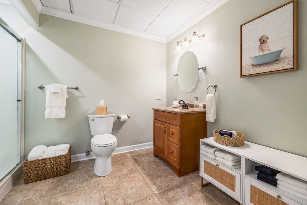 A light green bathroom featuring a wooden vanity with a round mirror, a white toilet, and a glass-enclosed shower, all set on light brown tiled floors. A framed picture of a dog in a tub adds a whimsical touch.