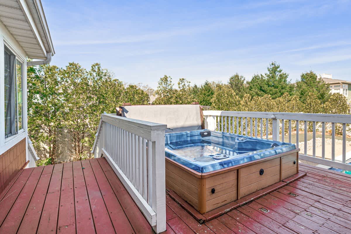 Outdoor hot tub with a wooden base and blue interior, situated on a red-stained wooden deck with white railings, surrounded by green trees under a bright blue sky.