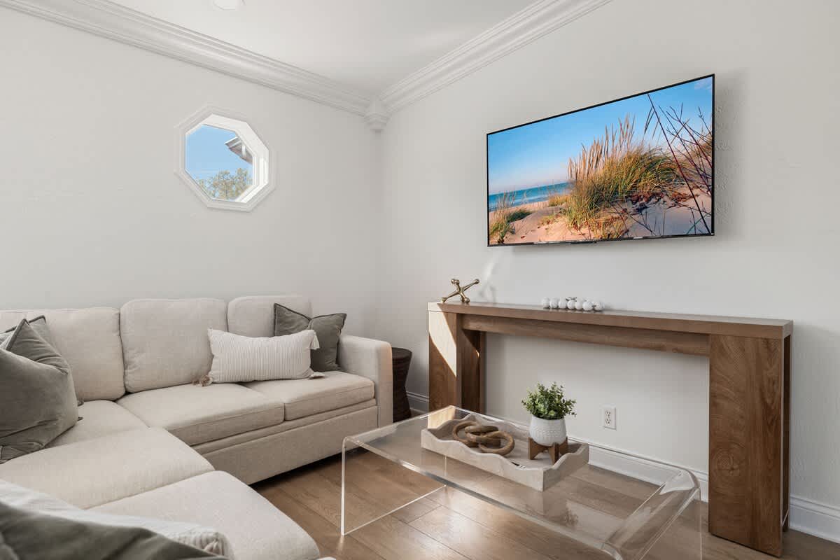 A bright living room with light wood floors, featuring a neutral L-shaped sectional sofa and a wall-mounted flat-screen TV displaying a beach scene. A clear acrylic coffee table and a wooden console table are also present.
