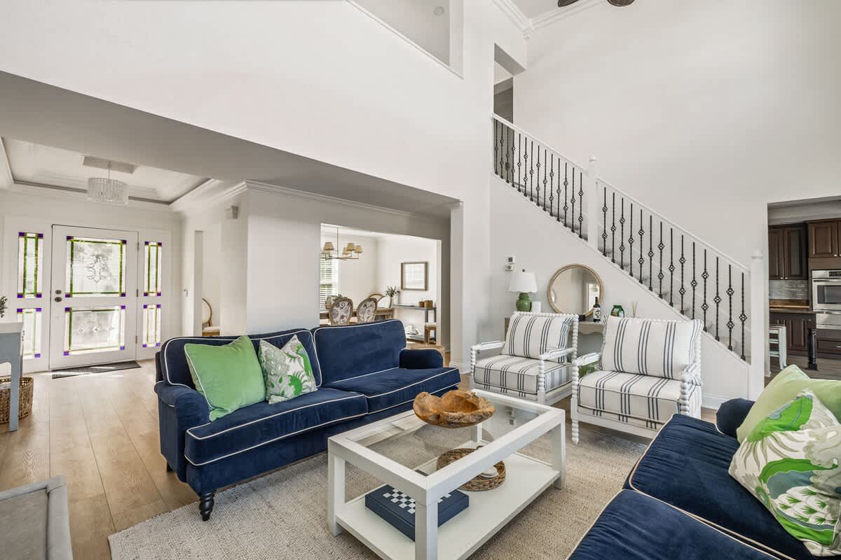 Bright and airy living room featuring a navy blue sofa with white piping, two striped armchairs, a glass-top coffee table, and a grand staircase with wrought iron railings, all set on light wood flooring.