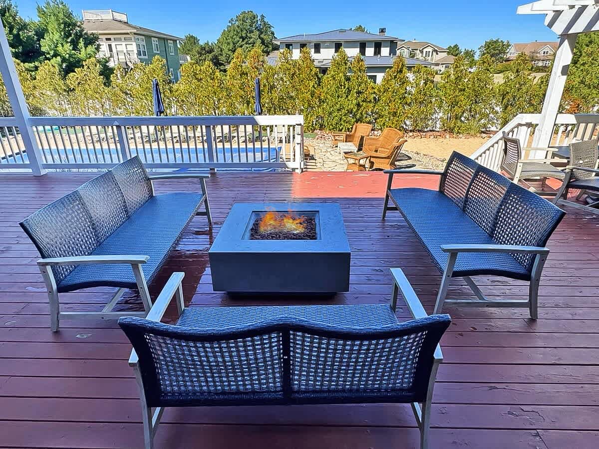 A cozy outdoor seating area on a wooden deck, centered around a rectangular concrete fire pit with a flickering flame. Two long benches and a smaller bench with dark woven backs provide ample seating for gathering.