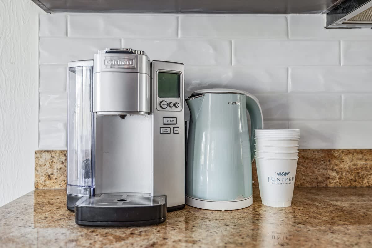 Close-up of a kitchen counter featuring a silver Cuisinart coffee maker, a light blue electric kettle, and disposable cups, set against a white subway tile backsplash and granite countertop.