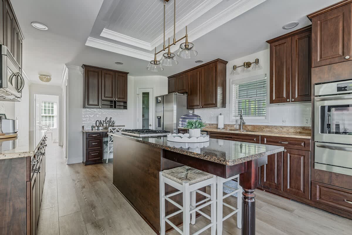 Modern kitchen featuring dark wood cabinetry, granite countertops, stainless steel appliances, a large island with a gas cooktop, and light wood flooring, with a breakfast nook visible in the background.