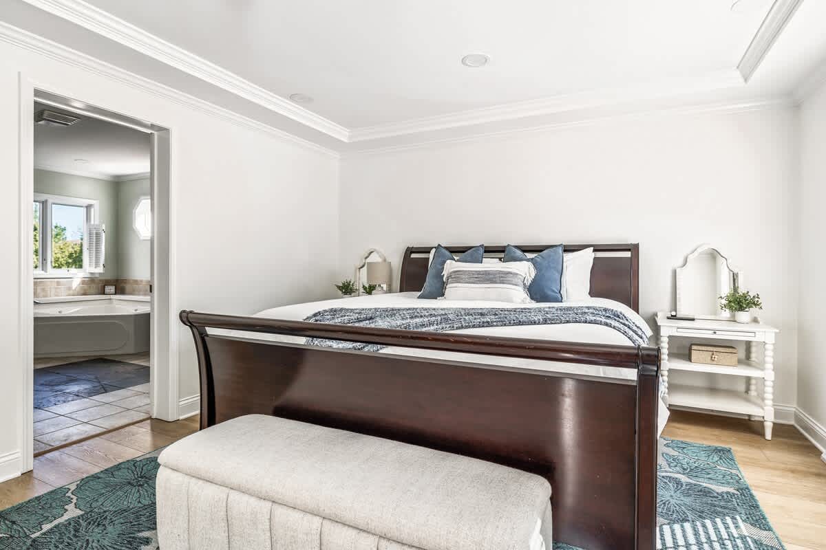 A spacious bedroom featuring a dark wood sleigh bed with white bedding and blue accent pillows, complemented by a light grey upholstered bench at the foot. A blue and white patterned rug covers the light wood floor, and a bathroom is visible through an open doorway.