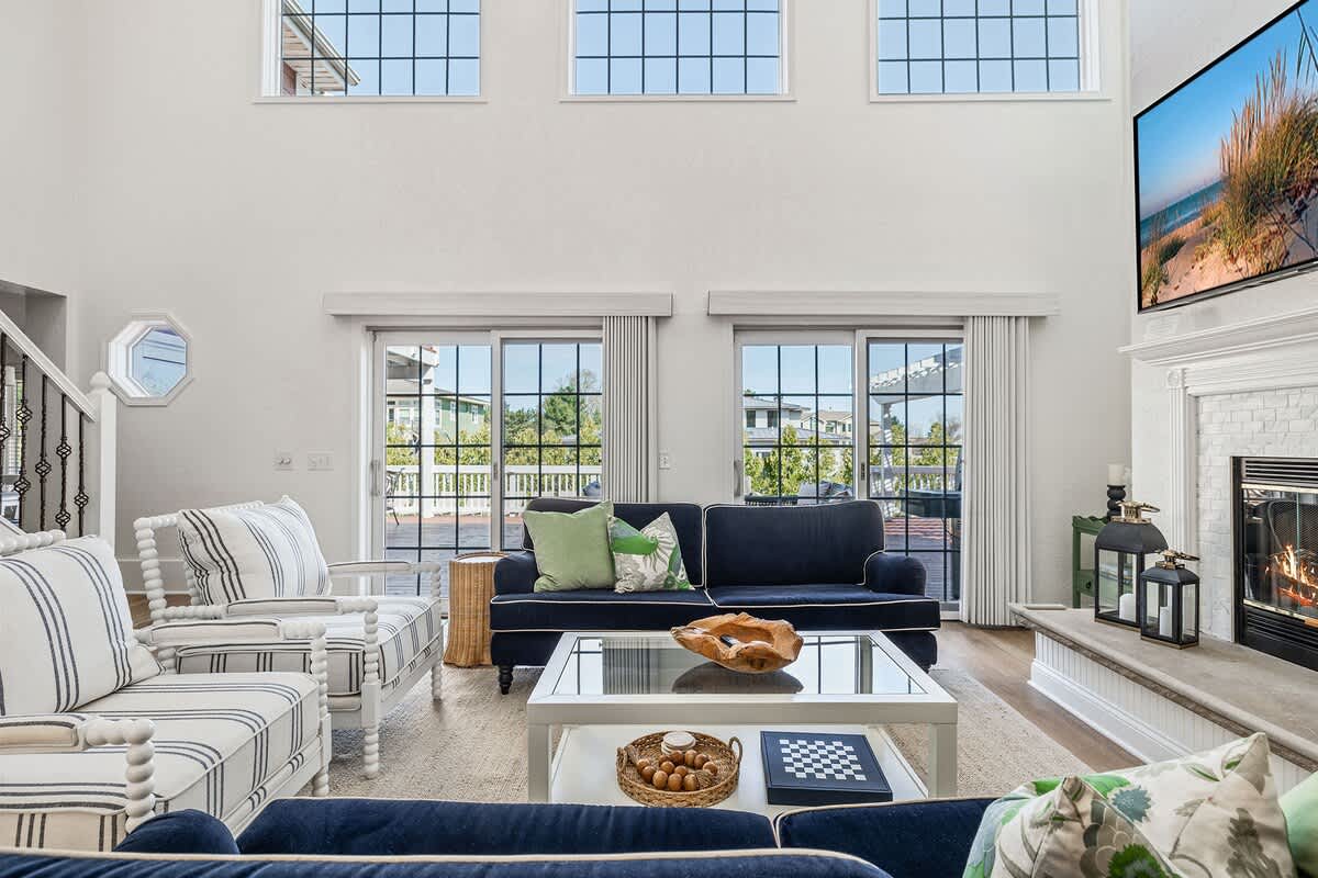 A bright living room featuring high ceilings and large sliding glass doors that open to a deck. The space is furnished with dark blue sofas and striped armchairs, centered around a glass coffee table.
