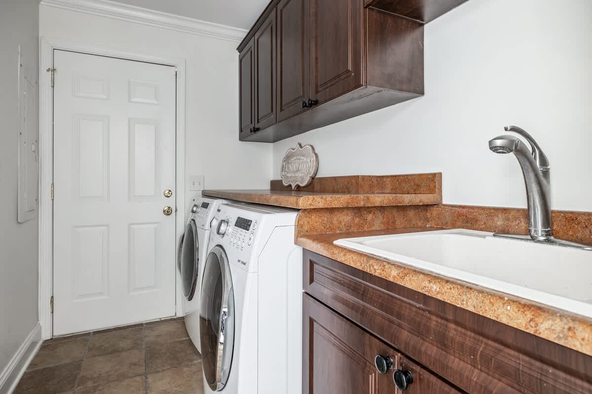 A functional laundry room featuring a white washer and dryer set, a light brown speckled countertop with a sink and chrome faucet, and dark wood upper and lower cabinets. A white paneled door is visible on the left.