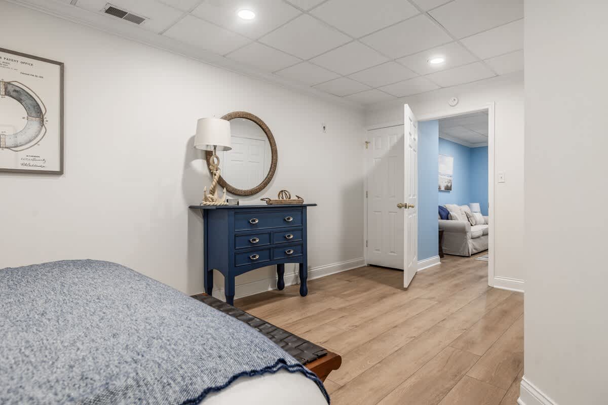 View of a bedroom with white walls, a dark blue dresser with a round mirror above it, and a wooden floor. A doorway leads to another room with blue walls, suggesting an adjacent living space.