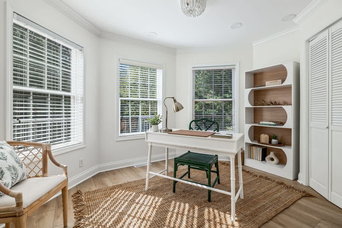 Bright home office with light wood floors, a white desk, and a green bamboo-style chair. Multiple windows with blinds provide ample natural light, and a white and wood bookshelf stands against the wall.