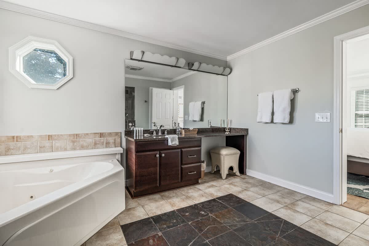 A wide shot of a bathroom with light grey walls, showcasing a jetted tub, a dark wood double vanity with a granite top, and a small stool. The floor features large brown and dark grey tiles, and an octagonal window is visible.