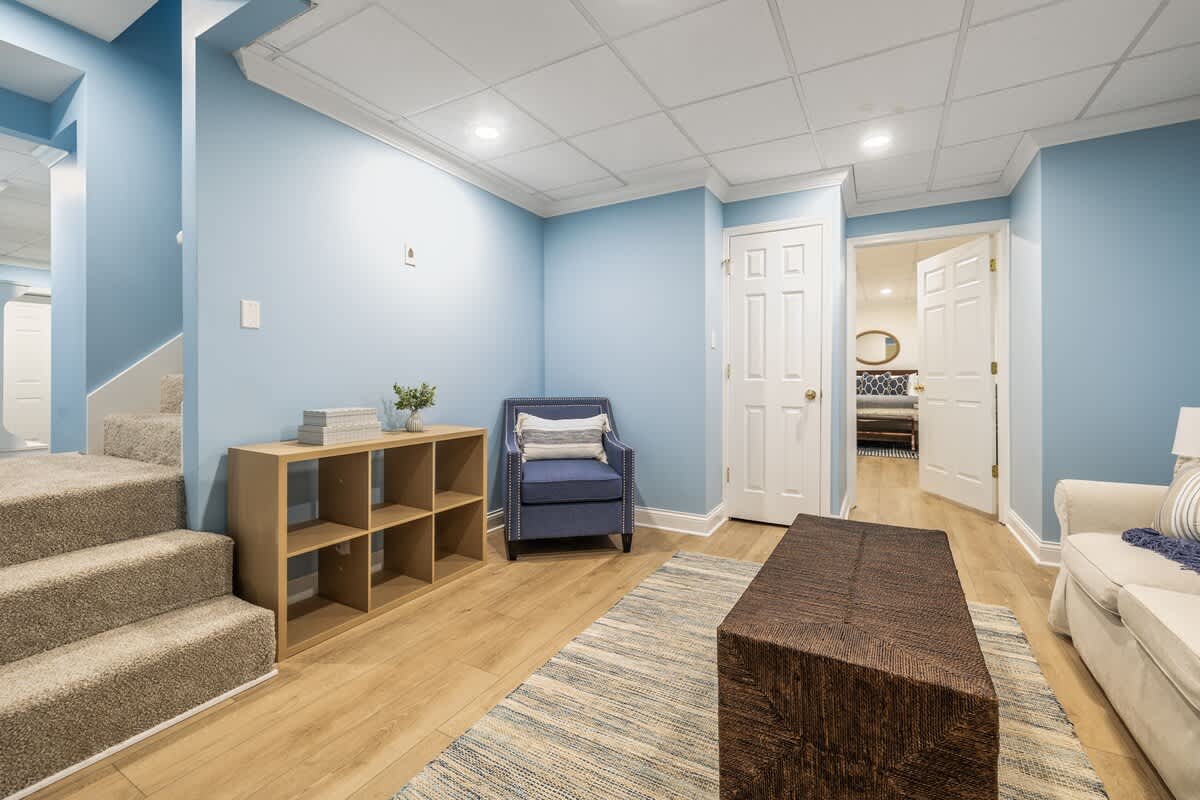 Cozy living area with light blue walls, light wood floors, and a drop ceiling. A blue armchair and a wooden cube shelf are visible, with carpeted stairs leading up on the left.