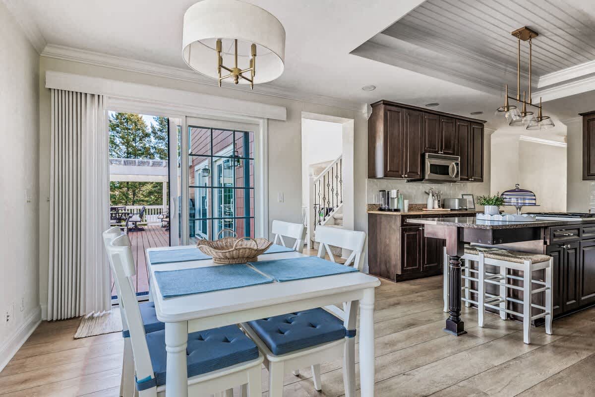 Bright dining area with a white rectangular table set with blue placemats, surrounded by white chairs with blue cushions, next to a kitchen with dark wood cabinets and a large sliding glass door leading to a deck.