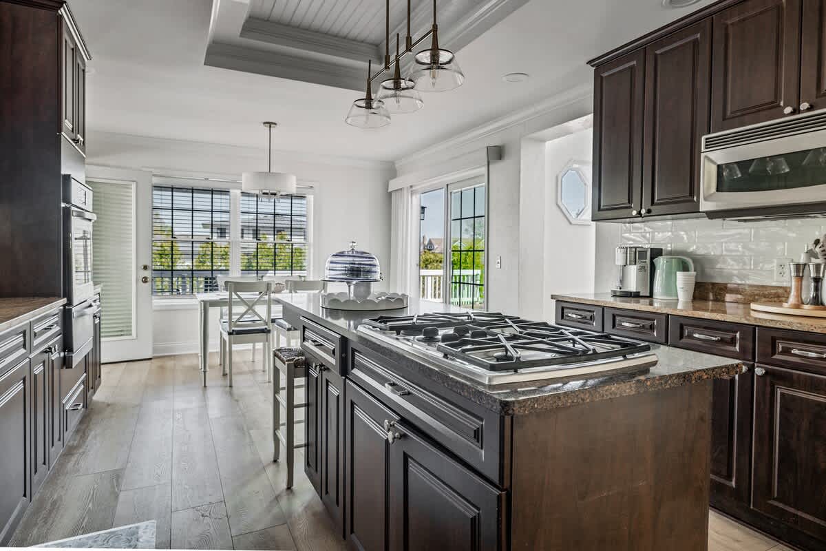 Well-appointed kitchen with dark wood cabinetry and a central island, leading to a breakfast area with a white table and chairs, illuminated by natural light from large windows and sliding glass doors.