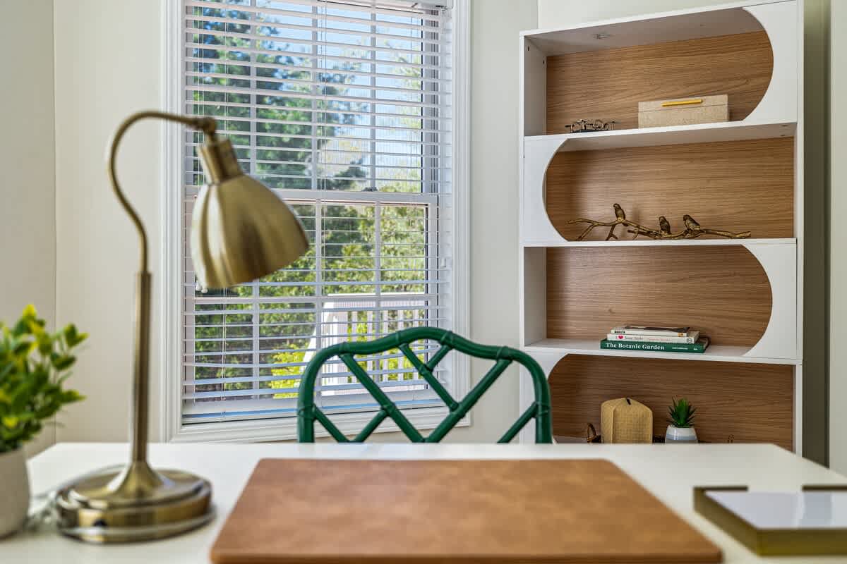 Close-up of a white desk with a gold task lamp and a brown desk pad, positioned in front of a window with blinds. A green bamboo-style chair is tucked under the desk, and a white and wood bookshelf is visible to the right.