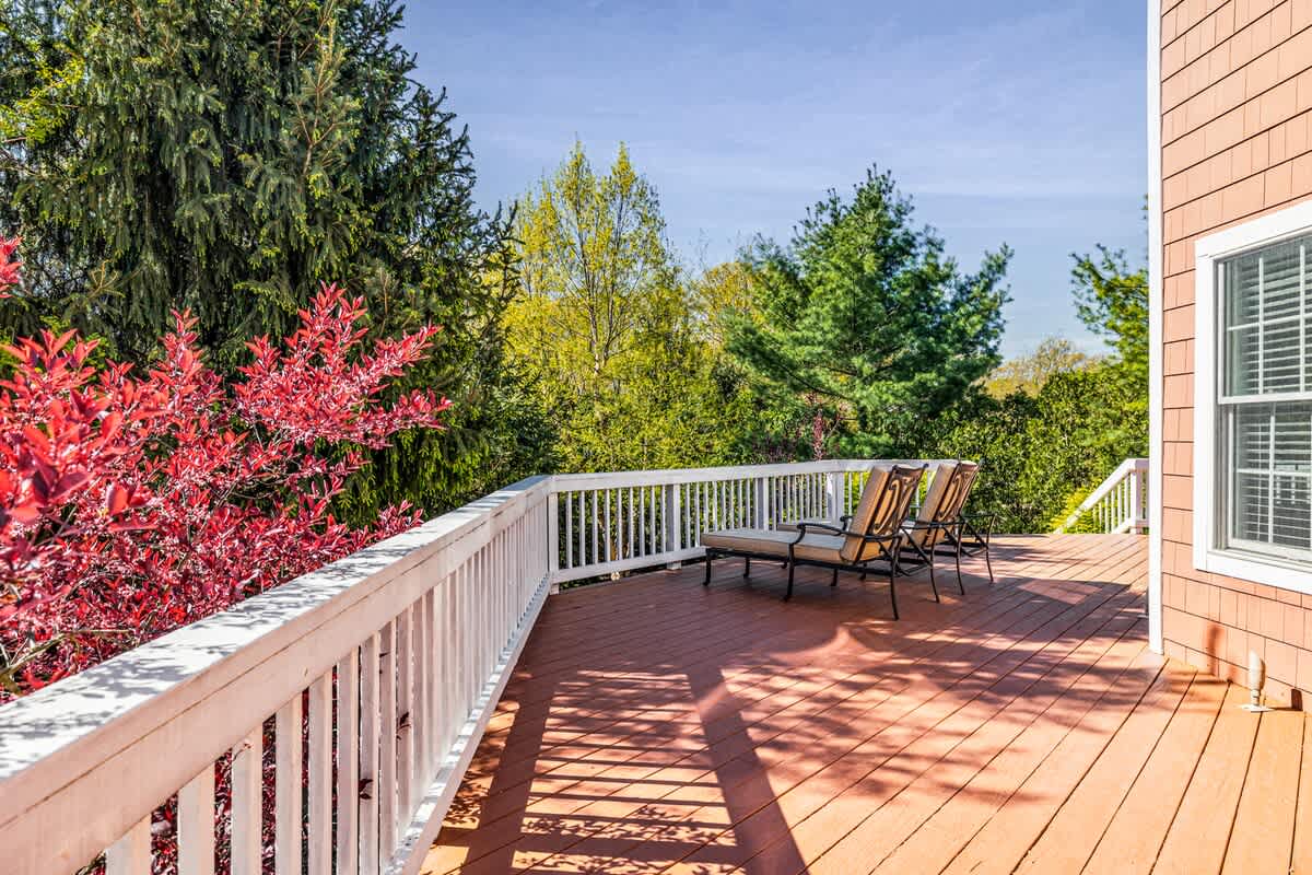 A sunny wooden deck with white railings, offering a serene outdoor space with two lounge chairs. The deck is surrounded by vibrant green trees and a striking red-leafed bush, providing a natural and private setting.