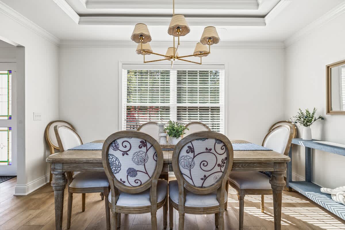 Elegant dining room with a rustic wooden table, six upholstered chairs featuring floral patterns, a brass chandelier with fabric shades, and a large window with white blinds, all on light wood flooring.