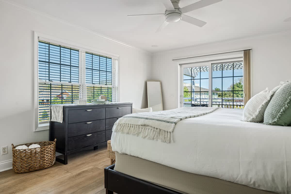 Bright bedroom with a white bed, dark wood dresser, and light wood floors. A large window with blinds and a sliding glass door offer natural light and access to an outdoor space.