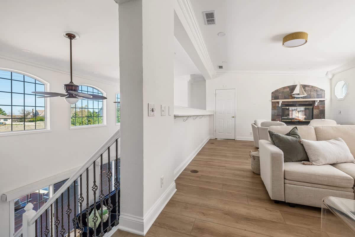 An upstairs landing with light wood flooring and a white railing with dark metal balusters, leading into a bright living area. Large arched windows offer views, and a ceiling fan is visible.