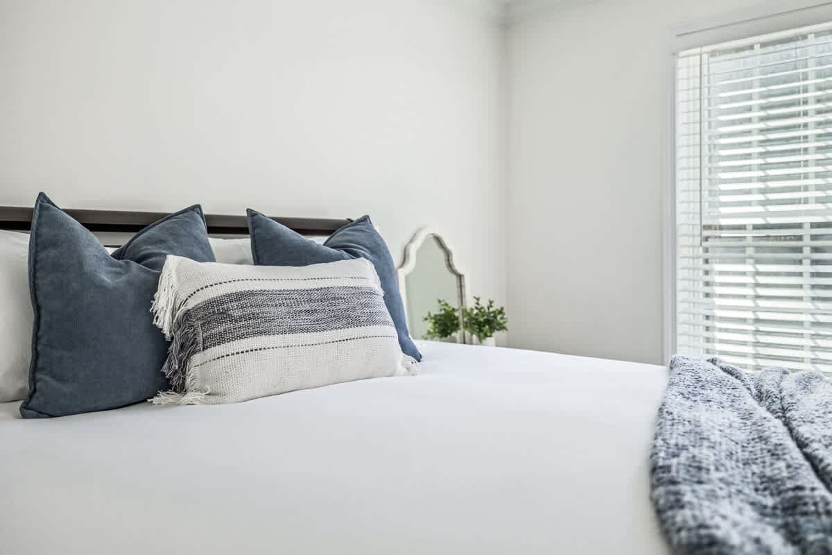 A close-up of a bed with crisp white bedding, dark blue pillows, and a white and grey striped decorative pillow. A dark headboard is visible, and a window with horizontal blinds is in the background.