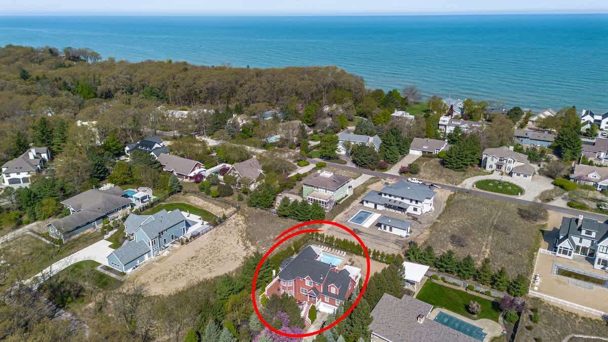 A wide aerial shot highlighting the property, circled in red, showing its location relative to a large body of water (likely a lake or ocean) in the background. The house is nestled among other residential properties and abundant greenery.