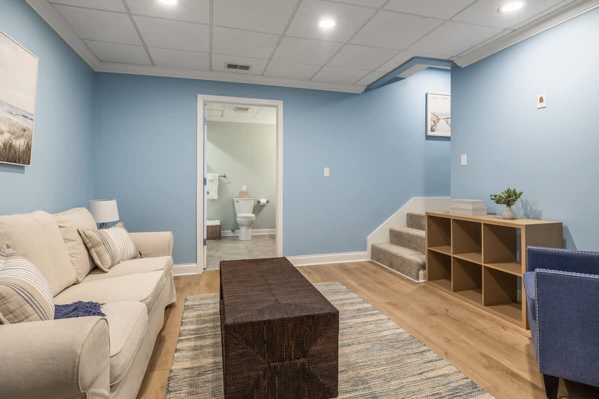 Spacious living area with light blue walls, light wood floors, and a beige sofa. A dark woven coffee table and a striped rug anchor the space, with a doorway leading to a bathroom in the background.